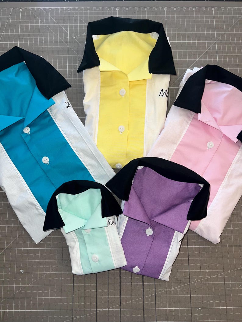 Op de afbeelding: Vijf kleurrijke bowlinghemden in teal, geel, roze, mintgroen en paars. Elk shirt heeft een wit middenpaneel, een zwarte kraag en witte knopen. Sommige shirts hebben handgeschreven namen.