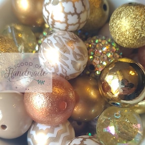 May include: A collection of gold, white, and copper colored beads with various patterns and textures. The beads are arranged in a bowl and include geometric, animal print, and solid colors.