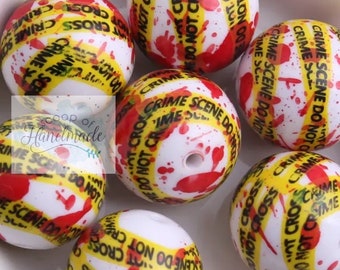 May include: Close-up of several white round beads with a red and yellow crime scene tape design. The beads have a glossy finish and are covered in red splatter paint.