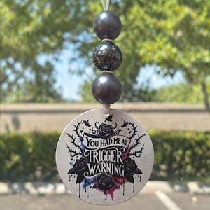 May include: A car air freshener with a round, white pendant featuring the text "You Had Me At Trigger Warning" surrounded by black roses and thorns. The pendant hangs from three dark gray beads.