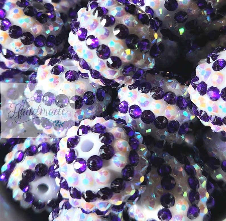 May include: Close-up of white iridescent round beads with purple rhinestones. The beads are clustered together and have a sparkly finish.