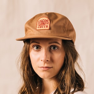 Peut inclure: Une femme portant une casquette de baseball marron avec un patch rouge et jaune qui dit "Tacos & Beer".