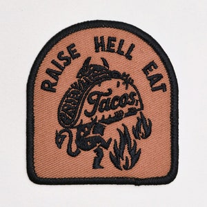 Może przedstawiać: Brązowa naszywka haftowana czarnym ściegiem. Naszywka przedstawia kreskówkowego diabła trzymającego taco z tekstem "Raise Hell Eat Tacos" wokół obrazu.