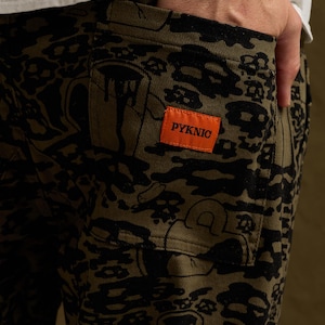 May include: Olive green pants featuring a black graphic print of various shapes and a rectangular orange patch on the back pocket with the text PYKNIC printed in black. The pants have a visible back pocket and a sturdy fabric construction.
