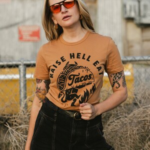 May include: A woman wearing a brown t-shirt with a black graphic that says "Raise Hell Eat Tacos" and black jeans. She is wearing orange sunglasses and has a tattoo on her arm.