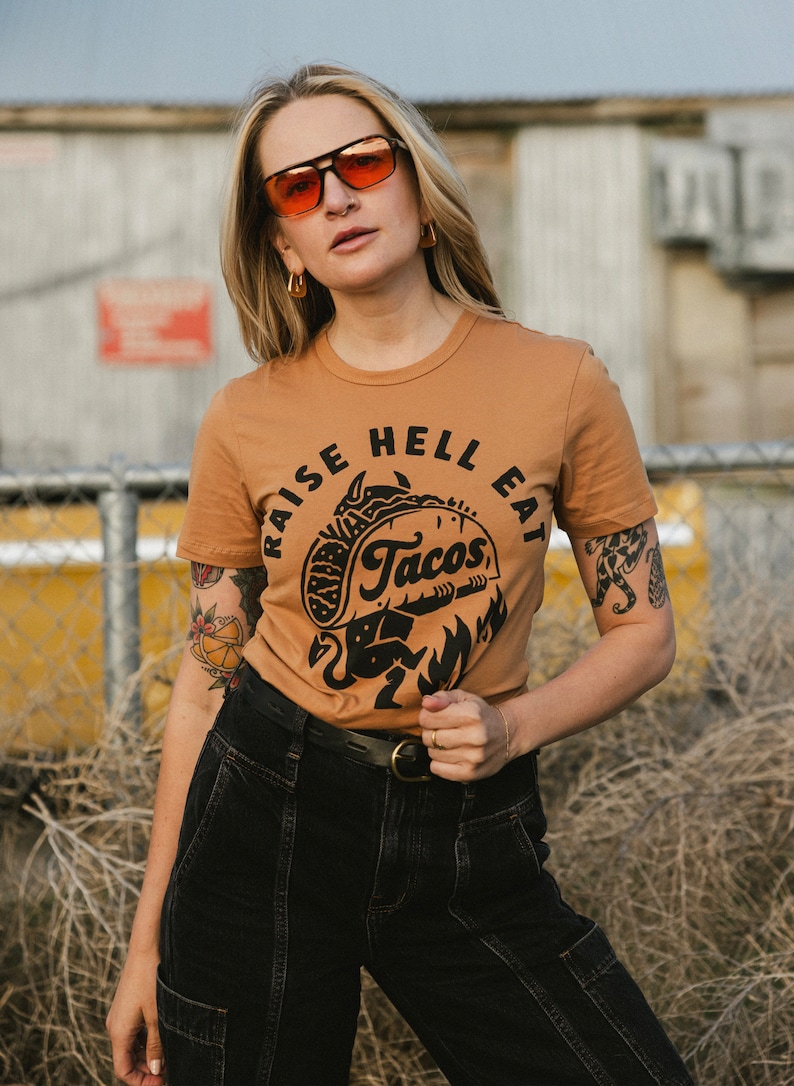May include: A woman wearing a brown t-shirt with a black graphic that says "Raise Hell Eat Tacos" and black jeans. She is wearing orange sunglasses and has a tattoo on her arm.