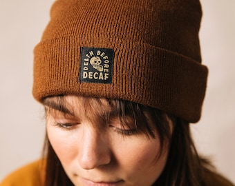 May include: A brown beanie with a folded brim. A black square patch on the front displays the words "DEATH BEFORE DECAF" around a skull graphic. The beanie is made of a ribbed knit material.