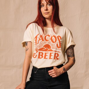 May include: A woman wearing a cream coloured t-shirt with the text "Tacos & Beer" in orange. The shirt features a graphic of a sunset and mountains. She is wearing black jeans.