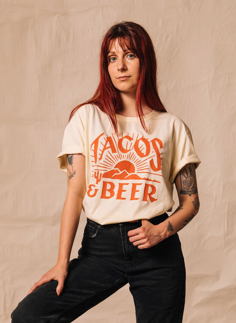 May include: A woman wearing a cream coloured t-shirt with the text "Tacos & Beer" in orange. The shirt features a graphic of a sunset and mountains. She is wearing black jeans.