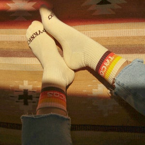 May include: Cream-colored socks with ribbed texture and the word "PYKNIC" printed on the toes. The cuffs feature a retro design with the word "TACOS" in a colorful stripe. The socks are worn with blue denim jeans.