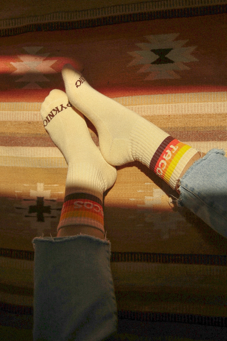 May include: Cream-colored socks with ribbed texture and the word "PYKNIC" printed on the toes. The cuffs feature a retro design with the word "TACOS" in a colorful stripe. The socks are worn with blue denim jeans.