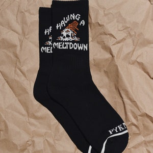 May include: A pair of black socks with a white skull graphic and the text "Having a meltdown". The socks have a white stripe at the top and the word "PYK" is visible on the bottom of one sock.