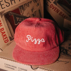 May include: A red corduroy baseball cap with the word "Pizza" embroidered in white script. The cap sits in front of pizza boxes with the words "Fresh Pizza" and a list of toppings.