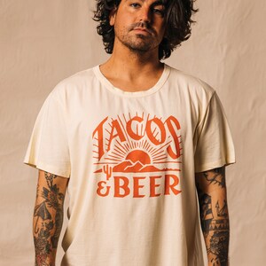 May include: A person wearing a cream coloured t-shirt with a red graphic that says "Tacos & Beer" with a sunset and mountains in the design.