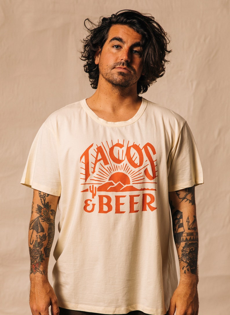 May include: A person wearing a cream coloured t-shirt with a red graphic that says "Tacos & Beer" with a sunset and mountains in the design.