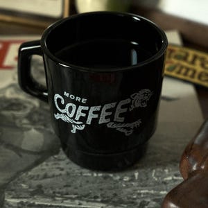 Könnte beinhalten: Schwarze Kaffeetasse mit quadratischem Griff. Auf der Tasse steht in weißer Schrift "MORE COFFEE" und eine Tiger-Illustration. Die Tasse steht auf einer gemusterten Oberfläche.