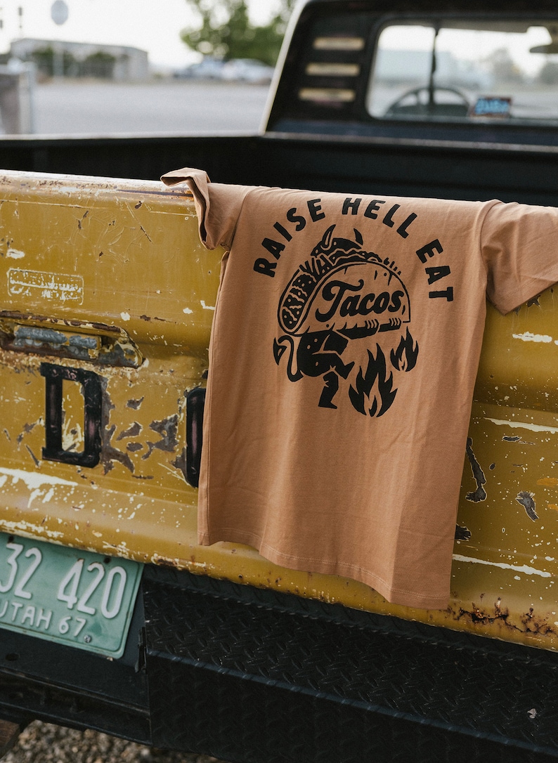 May include: A brown t-shirt with a black graphic of a cartoon devil holding a taco with the text "Raise Hell Eat Tacos".