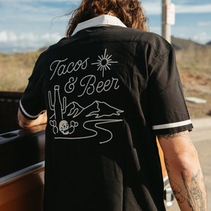 May include: Black bowling shirt with white embroidered text that reads "Tacos & Beer" and a desert scene with a cactus, a skull, and a sun.