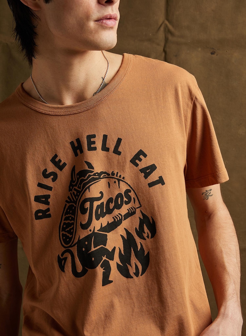 May include: A brown t-shirt with a black graphic print of a taco with flames and the text "Raise Hell Eat Tacos".