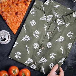 May include: Olive green button-down shirt with a white pizza, skull, and dagger pattern. A slice of pizza, tomatoes, and a can of soda are arranged around the shirt on a dark surface. The shirt has the text "Nutrition Facts by PYKNIO".