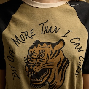Könnte beinhalten: Olivgrünes Raglan-T-Shirt mit schwarzen Ärmeln und einem cremefarbenen Ausschnitt. Das Shirt zeigt eine Tigergrafik und den Text "But Off More Than I Can Chew" in schwarzer Schrift, ein lässiges und kühnes Design.