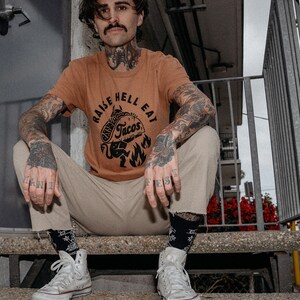 May include: A person wearing a brown t-shirt with the text "Raise Hell Eat Tacos" and a black and white illustration of a devil holding a taco. The person is sitting on a set of concrete steps wearing white high-top trainers.