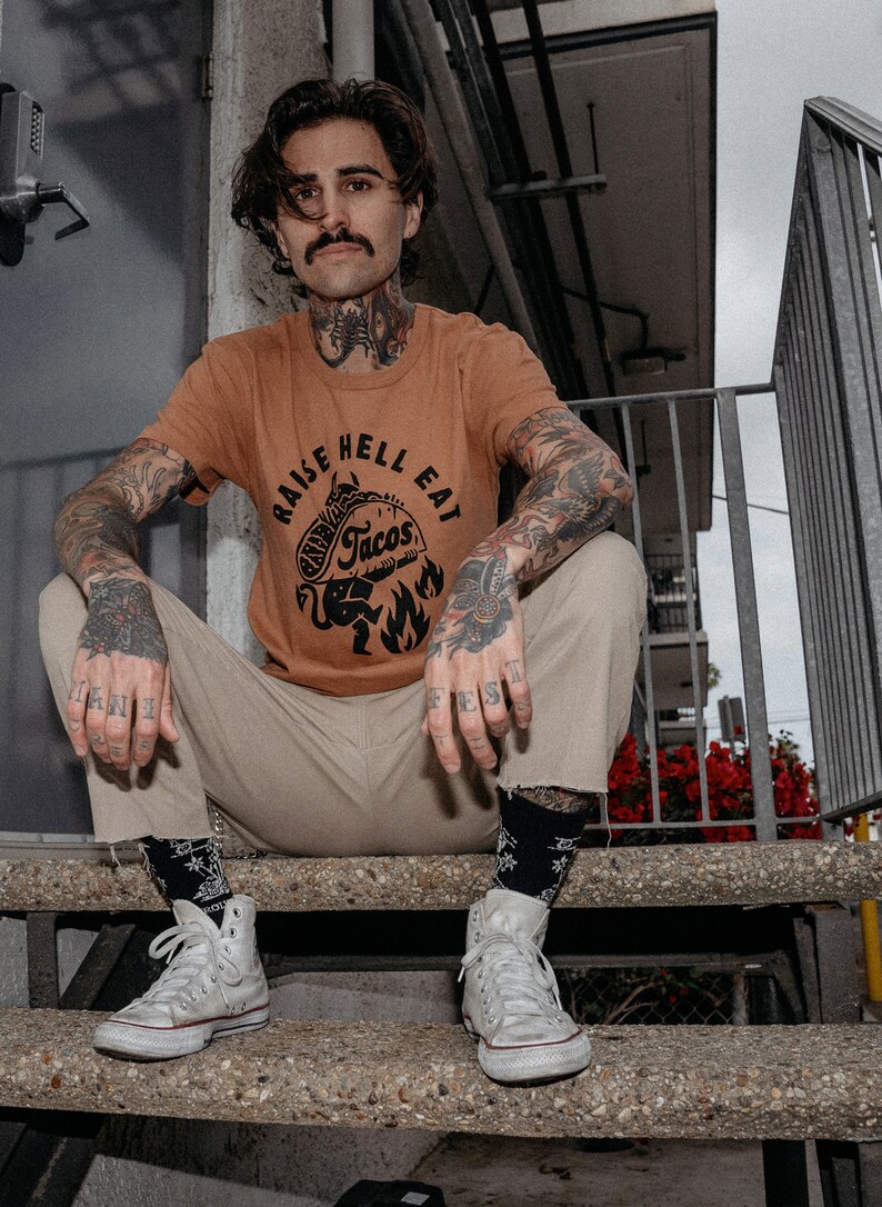 May include: A person wearing a brown t-shirt with the text "Raise Hell Eat Tacos" and a black and white illustration of a devil holding a taco. The person is sitting on a set of concrete steps wearing white high-top trainers.