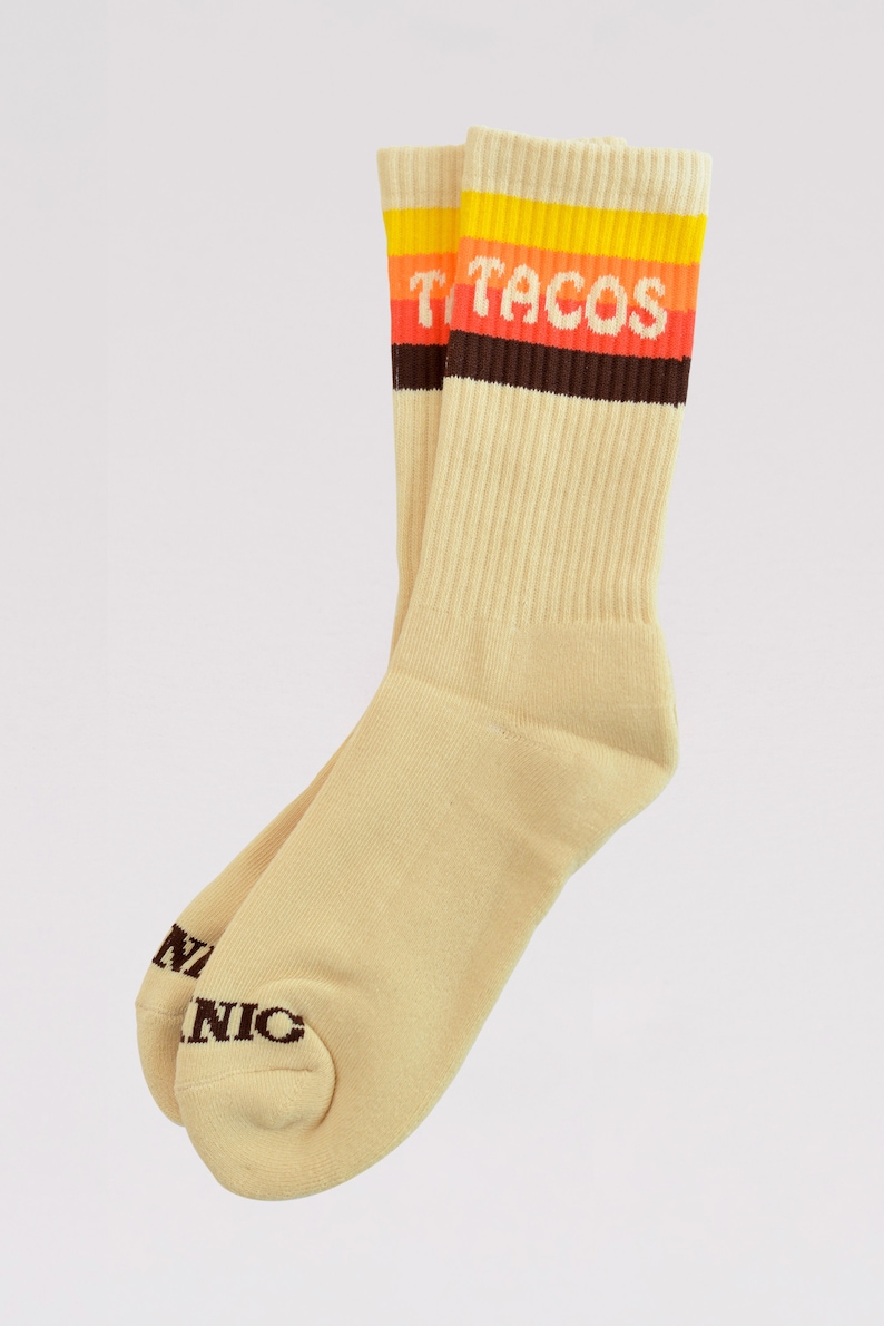 May include: A pair of beige crew socks with a ribbed design. The top band features the word "TACOS" in orange and white lettering, with yellow and brown stripes. The bottom of the socks has the word "NIC" in brown.