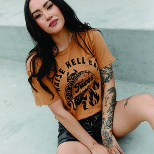 May include: A woman wearing a brown t-shirt with the text "Raise Hell Eat Tacos" and a black and white illustration of a taco with a devil's tail. She is wearing black shorts and has tattoos on her arms and legs.