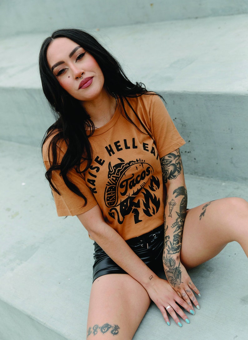 May include: A woman wearing a brown t-shirt with the text "Raise Hell Eat Tacos" and a black and white illustration of a taco with a devil's tail. She is wearing black shorts and has tattoos on her arms and legs.