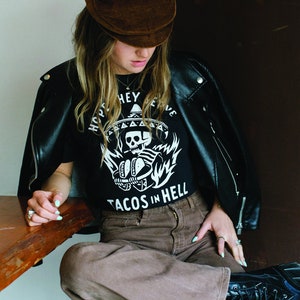 Peut inclure: Une personne portant une veste en cuir noire, un t-shirt noir avec un motif de cr&acirc;ne et d'os crois&eacute;s blancs et le texte "Hope They Serve Tacos In Hell", une casquette en velours c&ocirc;tel&eacute; marron et un pantalon marron.