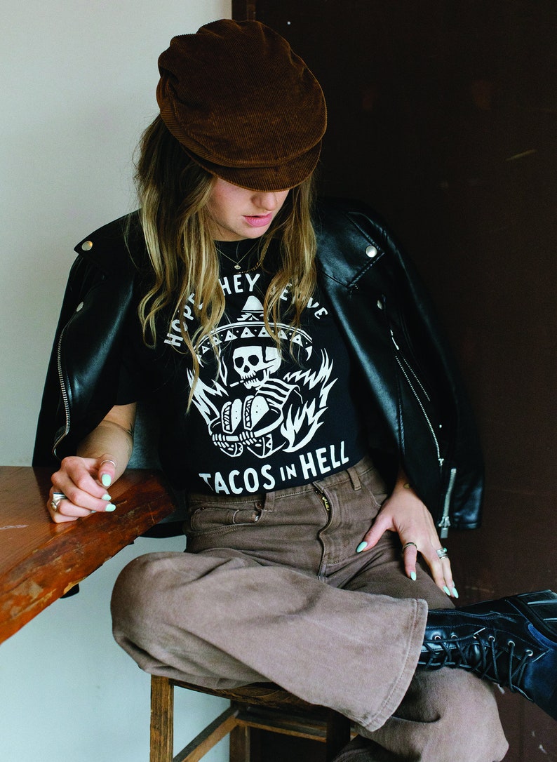 Peut inclure: Une personne portant une veste en cuir noire, un t-shirt noir avec un motif de cr&acirc;ne et d'os crois&eacute;s blancs et le texte "Hope They Serve Tacos In Hell", une casquette en velours c&ocirc;tel&eacute; marron et un pantalon marron.