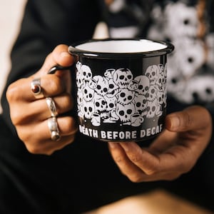 Könnte beinhalten: Schwarze Emaille-Tasse mit einem weißen Design aus Schädeln und dem Text "Death Before Decaf".