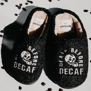 May include: A pair of black fuzzy slippers with white embroidery that reads "Before Decaf" and a skull graphic. The slippers are on a white background with coffee beans scattered around them.
