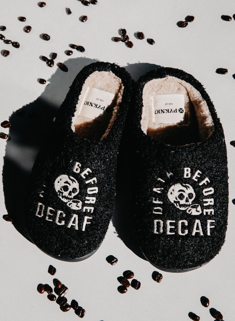 May include: A pair of black fuzzy slippers with white embroidery that reads "Before Decaf" and a skull graphic. The slippers are on a white background with coffee beans scattered around them.