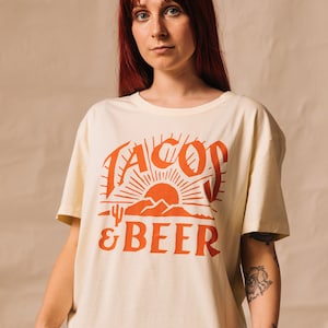 May include: A cream coloured t-shirt with a red graphic of a sunset, mountains, and a cactus with the text "Tacos & Beer".