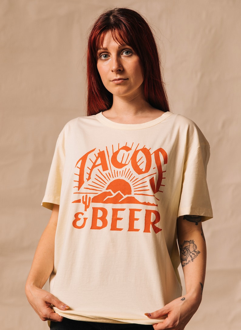 May include: A cream coloured t-shirt with a red graphic of a sunset, mountains, and a cactus with the text "Tacos & Beer".