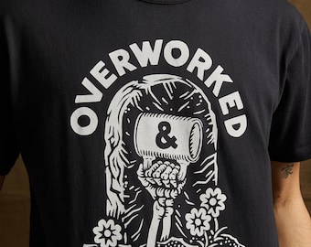 May include: Black t-shirt featuring a white graphic of a skeleton holding a coffee mug with the text "OVERWORKED & UNDER CAFFEINATED".