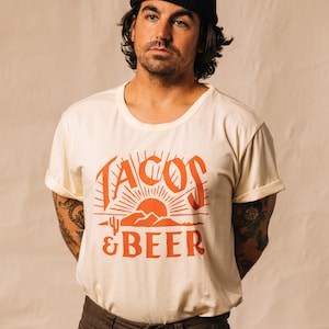 May include: A person wearing a white t-shirt with a red graphic that says "Tacos & Beer" and a black beanie. The graphic features a sunset with mountains and a cactus.
