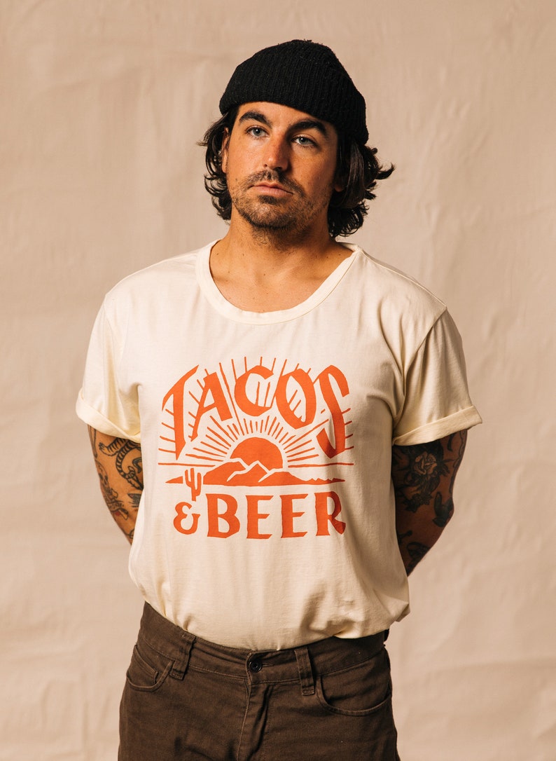 May include: A person wearing a white t-shirt with a red graphic that says "Tacos & Beer" and a black beanie. The graphic features a sunset with mountains and a cactus.