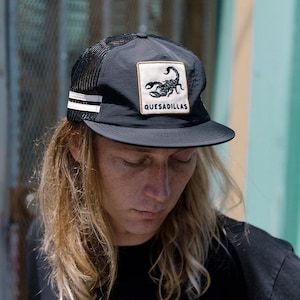 Può includere: Cappellino da camionista nero con retro in rete e visiera curva. Il cappellino presenta una toppa quadrata con un'immagine di scorpione e la scritta "QUESADILLAS". Due strisce bianche sono sul lato.
