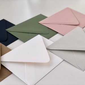 May include: A set of six envelopes in various colors, including brown, white, green, gray, pink, and navy blue. The envelopes are all folded and stacked on top of each other.
