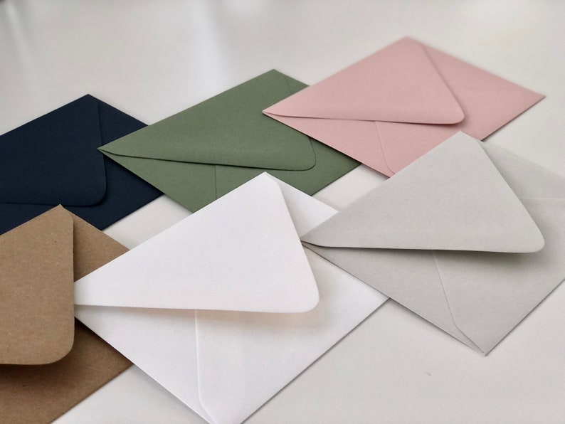 May include: A set of six envelopes in various colors, including brown, white, green, gray, pink, and navy blue. The envelopes are all folded and stacked on top of each other.