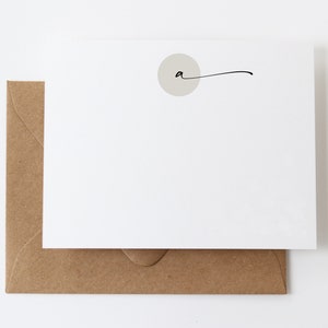 May include: A white stationery card with a light gray circle and a handwritten "a" with a curved line. The card is paired with a brown envelope. The stationery set is on a white background.
