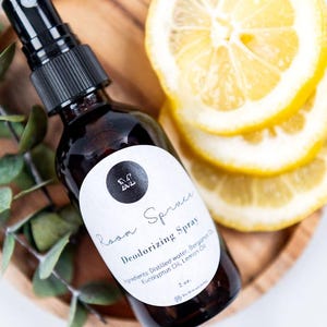 Op de afbeelding: Een 2 oz amberkleurige glazen fles met Room Spruce Deodorizing Spray en een zwarte spraykop. Het witte etiket bevat de letter M in een zwarte cirkel, de tekst Room Spruce Deodorizing Spray en een lijst met ingrediënten waaronder citroenolie.