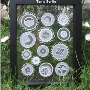 May include: A black framed picture with a glass front showcasing white lace circles of various designs. The title "Tanja Berlin" is printed at the top of the frame.