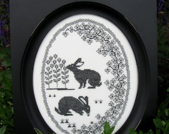 May include: Black and white cross-stitch embroidery of two rabbits in a garden setting, framed in a black oval frame.