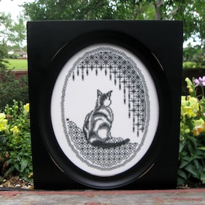 May include: Black and white embroidery of a cat sitting in a circle with a black and white lace design. The embroidery is framed in a black frame.