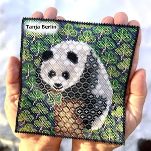 May include: A square beaded artwork featuring a panda bear in a forest setting. The panda is black and white, with a detailed pattern. The background is a mix of green and blue, with the text "Tanja Berlin" in the upper left corner.