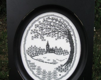 May include: Black and white embroidery of a tree with a house in the background, framed in a black oval frame.